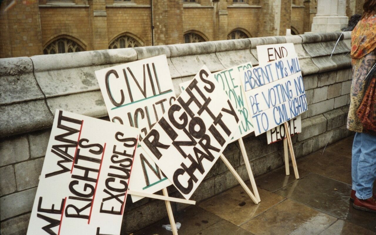 Six placards balanced against a stone wall with a person standing on the right edge of the frame. Most placards are overlapping, with some reading ‘We want rights not excuses’, ‘Civil rights now’, ‘Rights not charity’ and ‘Absent MPs will be voting no to civil rights’.