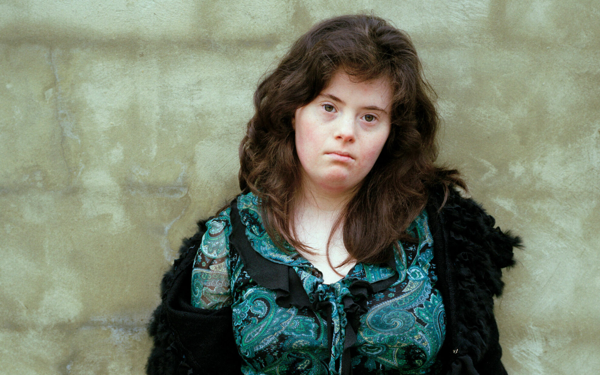 A young white woman with Down's Syndrome is stood against a plain stone wall staring directly at the camera. She has long dark hair and is wearing a patterned green and black blouse.