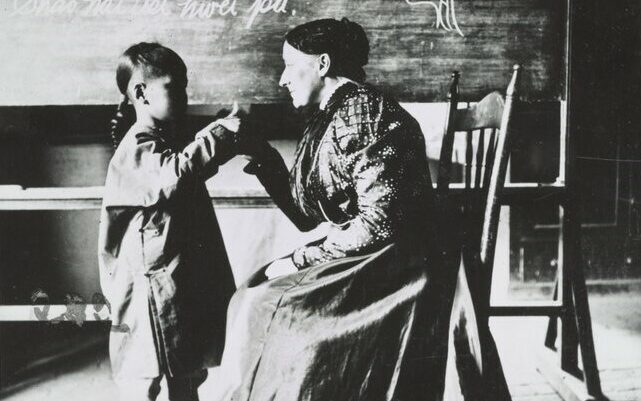 A black and white photograph of a white woman sat in a classroom facing a young Chinese boy stood in front of her. She is looking at him intently and is holding his hand aloft in a particular shape to teach him fingerspelling.