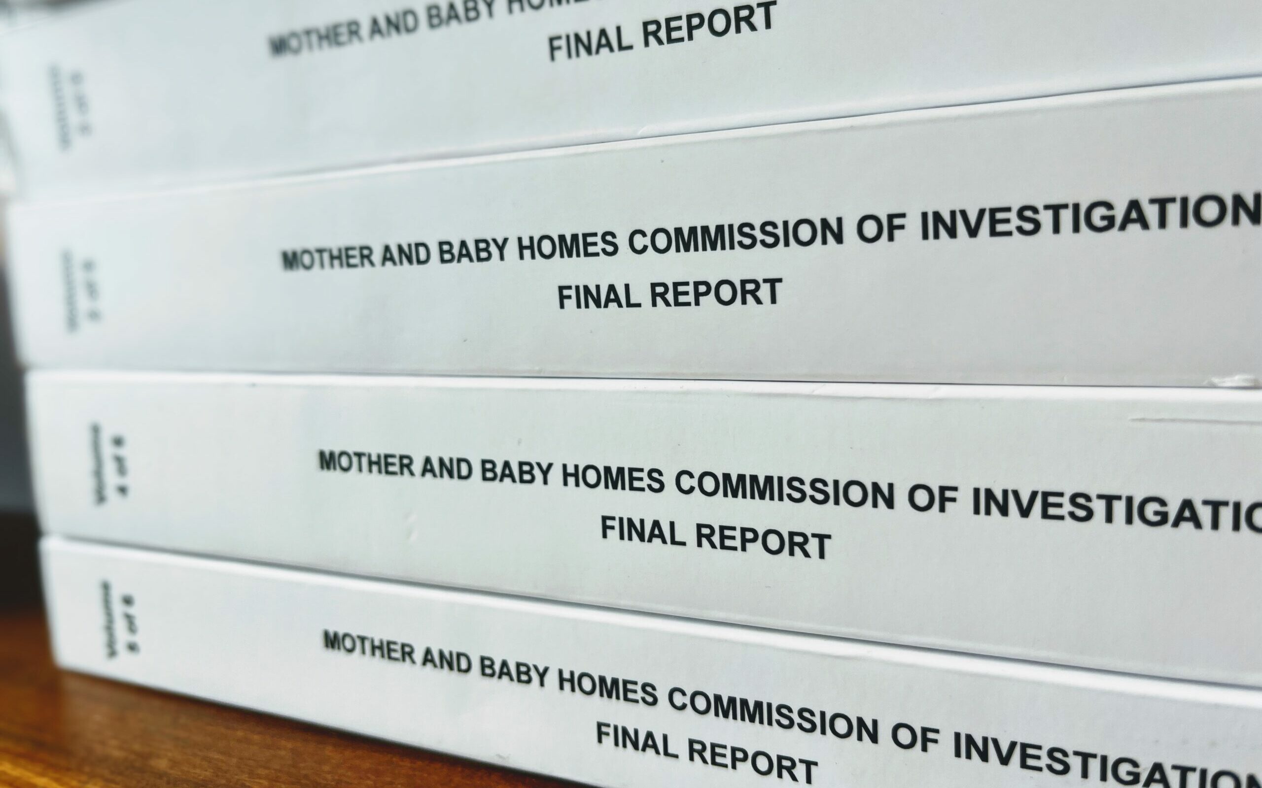 Four hardbound books stacked one on top of the other. The spine of each book is white and the title is printed in bold black letters 'Mother and Baby Homes Commission of Investigation: Final Report'.