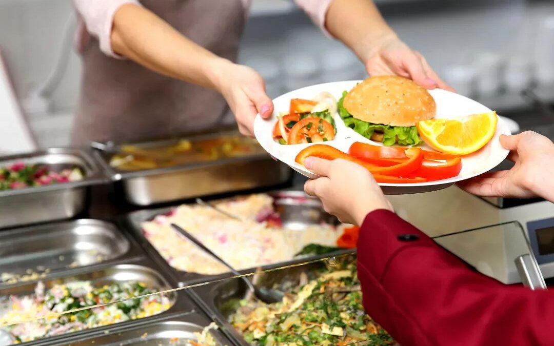 A colour photograph of the serving area of a school canteen. A child reaches for a tray being handed to them by a dinner lady. On their plate is a burger and some salads.