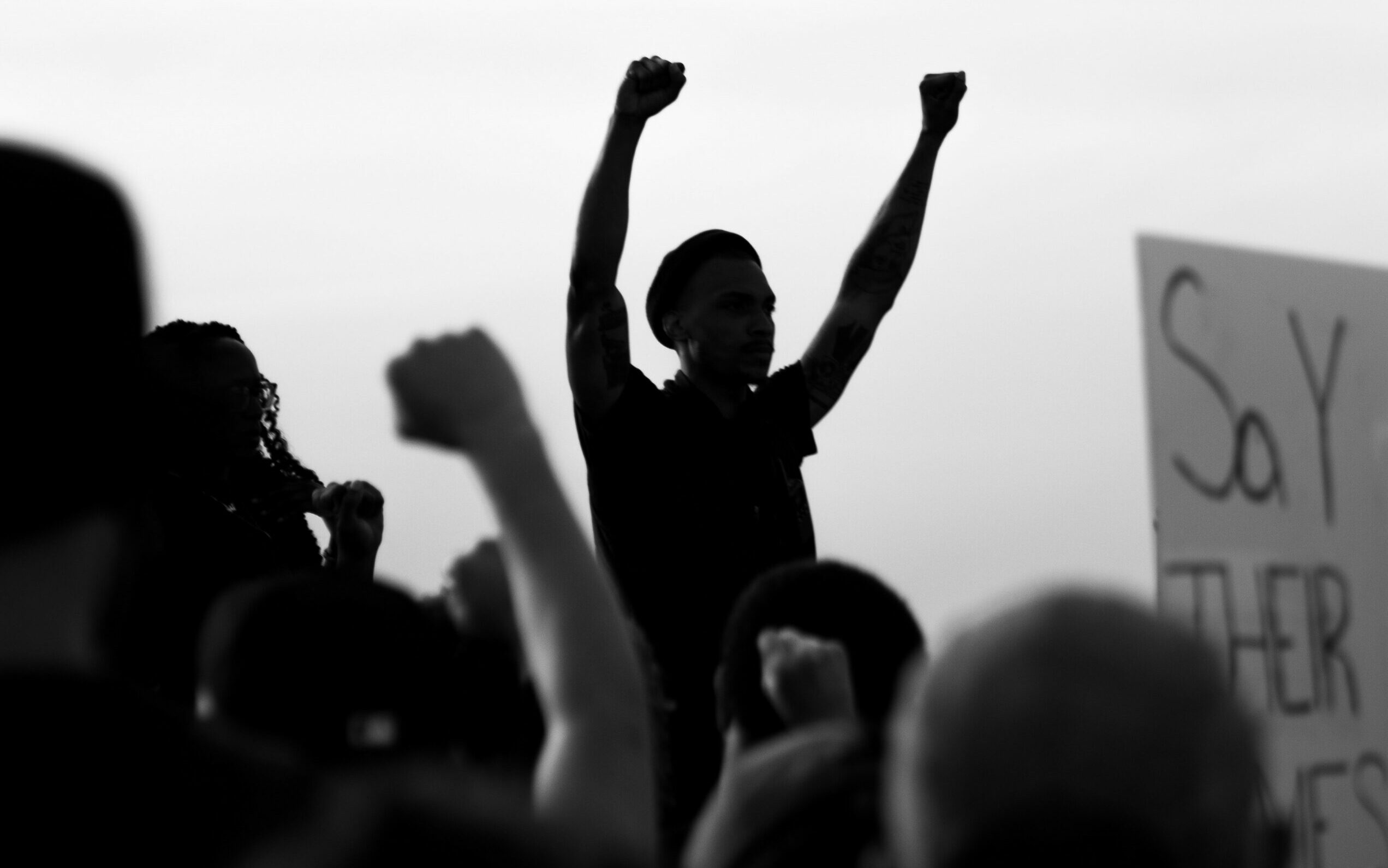 A black and white photograph of a crowd scene at a Black Lives Matter protest. In the foreground are people with their backs to the camera, several with raised fists. In the background is a striking silhouette of one person addressing the crowd with both their fists raised. A placard which reads 'Say Their Names' is just visible on the right.